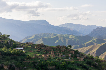 Fototapeta premium Wide landscape view of the abandoned stone village of Gamsutl on a green mountain ridge, with layered Caucasus peaks and clouds, Dagestan, Russia