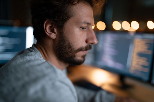Focused coder stares at glowing screens. Soft bokeh lights frame the quiet workspace. Deep concentration fills his thoughtful expression. Ideal for tech, development, or remote work themes
