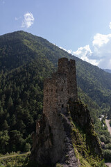 Naklejka premium Ancient Vovnushki tower complex perched on a rocky cliff above a green mountain valley in the Republic of Ingushetia, Russia, under blue sky with clouds