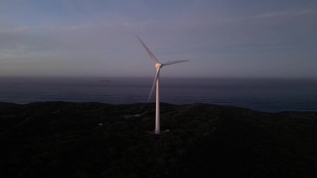Aerial drone shot of coastal wind turbine rotating above dark hillside terrain and ocean horizon during peaceful evening twilight atmosphere