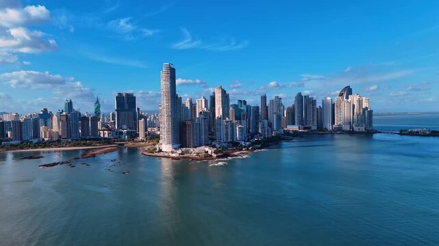 Punta Pacifica peninsula skyline rising from the Pacific coast, Panama City. Low angle drone view of towers on rocky point with calm ocean and wispy clouds.