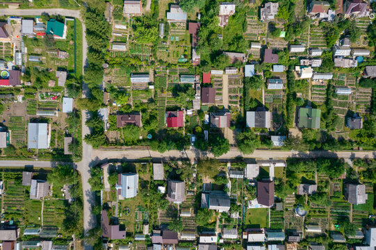 Top down aerial view of a suburban neighborhood with small houses, garden plots, greenhouses, and dirt roads surrounded by lush summer greenery