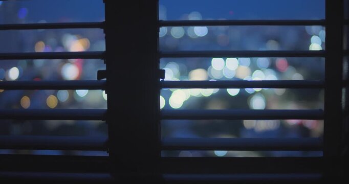 View of city at night from apartment or office building window with vertical blinds. Out of focus lights in background as window frame is in silhouette. Suspicious mysterious looking vibe in dark room