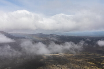 Fototapeta premium Wide view of mist and low clouds drifting over a dark volcanic plain and distant hills in Tolbachinsky Dol, Northern Breakthrough area, Kamchatka Krai, Russia