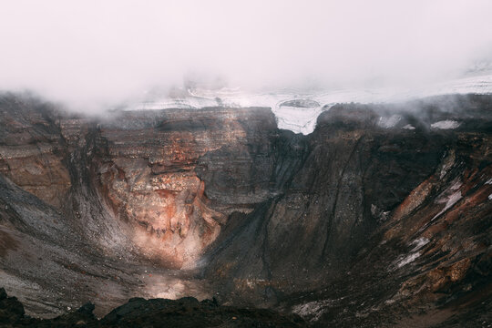 Dramatic view into the Tolbachik volcano crater with layered lava rock, snow patches and low clouds. Remote wilderness scenery in Kamchatka Krai, Russia