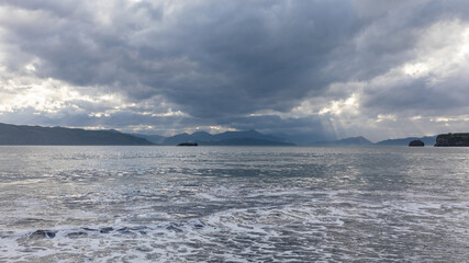 Fototapeta premium Dramatic cloudy sky and sunbeams over the cold sea with gentle surf and distant mountains, Avacha Bay, Kamchatka Krai, Russia, natural travel landscape
