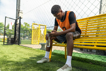 Young male soccer player sitting on yellow bench using smartphone during break, football athlete holding water bottle resting on turf field after sports training match outdoor © Supachai