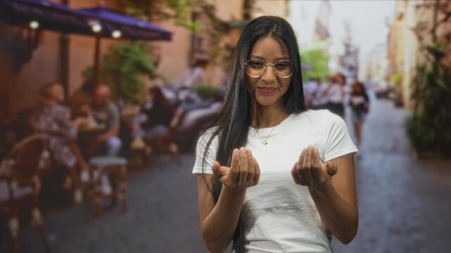 Woman showing palms and smiling, wearing glasses and a white t shirt in a street cafe with blurred diners and tables; friendly invitation.