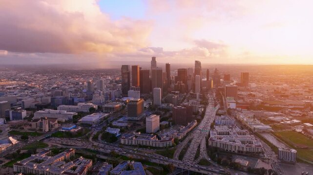Aerial view captures the downtown Los Angeles skyline at sunset. Traffic congestion fills the highways as commuters travel through the financial district. Palm trees outline the urban landscape.