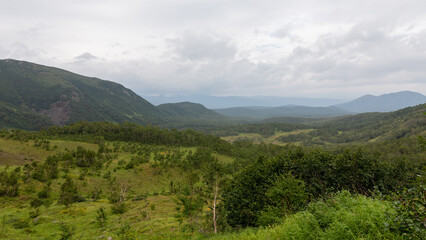 Obraz premium Wide scenic view of the Vachkazhets mountain range with lush green valley, forested hills and distant peaks under overcast clouds in Kamchatka Krai, Russia