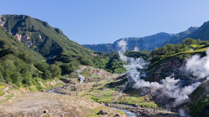 Wide view of the Valley of Geysers with steaming fumaroles, hot springs and a river in a green mountain valley, Kamchatka Krai, Russia, on a clear summer day © TravelFlow