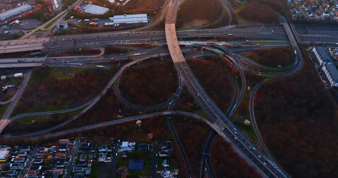 Aerial view of highway interchange at dusk surrounded by autumn trees. Overhead drone shot of multilane cloverleaf ramps, moving vehicles and residential buildings in warm fading light.