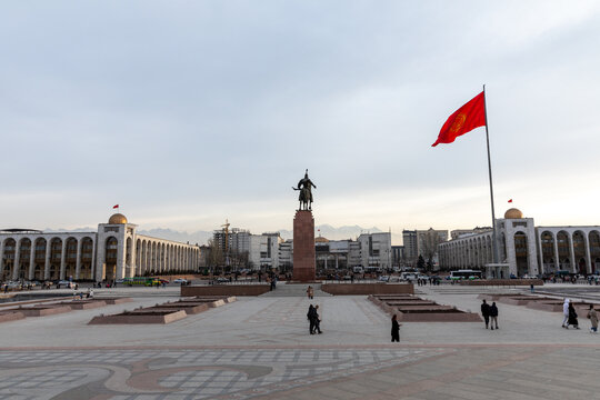 Bishkek, Kyrgyzstan - February 23, 2025: Wide view of Ala-Too Square with the Manas monument, government buildings and a large Kyrgyzstan flag, with pedestrians and mountains in the distance, Bishkek