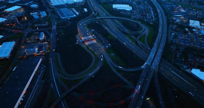 Overhead view of a highway toll plaza at dusk, United States. Top down vertical angle displaying multiple lanes, illuminated booths, adjacent parking lots, and deep evening shadows.