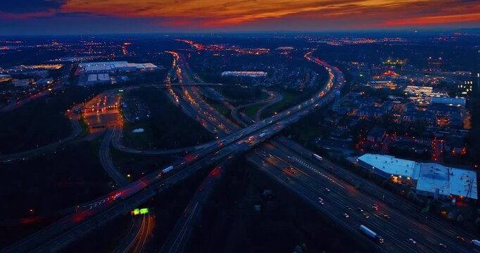 Elevated view of a highway toll plaza and traffic, United States. High wide angle shot featuring glowing collection booths, long lines of car lights, industrial buildings, and a dark horizon.