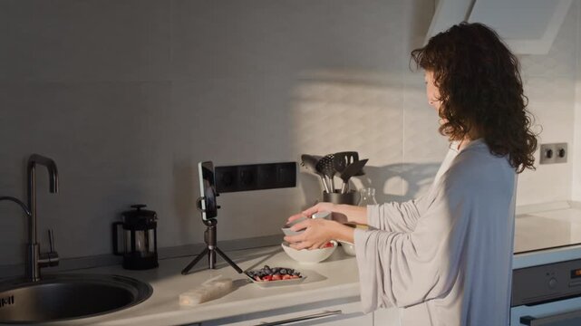 Young adult Caucasian woman setting up smartphone on tripod in kitchen, recording video blog about healthy breakfast and eating strawberry. Clip perfect for social media marketing campaigns