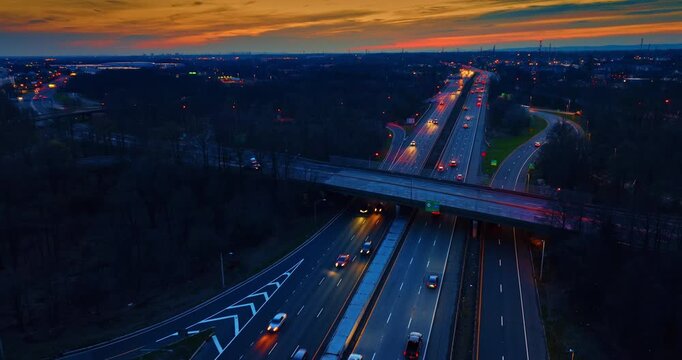 Highway traffic passing under an overpass bridge, United States. Elevated viewpoint showcasing blurred vehicle lights on multi lane asphalt, orange sunset sky on the horizon, and dark forest trees.