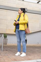 Mature adult woman standing on tiled plaza in yellow jacket, holding smartphone, wearing brown bag © wavebreak3