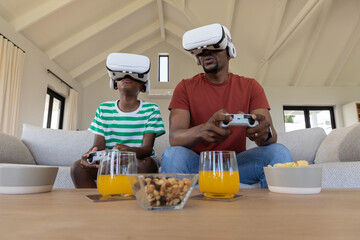 Father and son sitting on light sofa in living room wearing white VR headsets holding controllers © wavebreak3