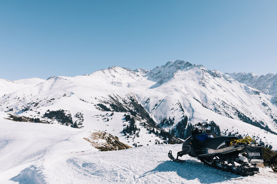 Snowmobile parked on a groomed snowy ridge with panoramic Tian Shan mountain peaks under clear blue sky at Karakol ski resort near Karakol, Issyk-Kul Region, Kyrgyzstan
