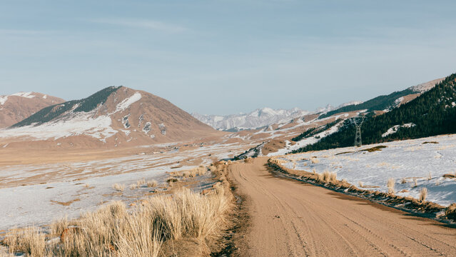 Scenic winding dirt road crossing a snowy mountain valley with dry grass and distant peaks, rural highlands of Kyrgyzstan in Central Asia, winter travel concept