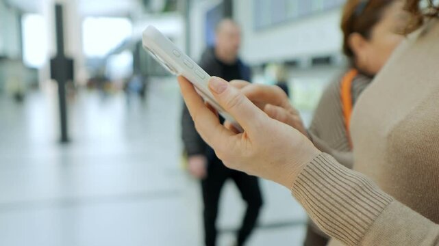 Person holds phone while walking through busy station. Blurred people move in background. Hands use device for navigation or communication. Indoor setting with bright lighting. Focus stays on phone