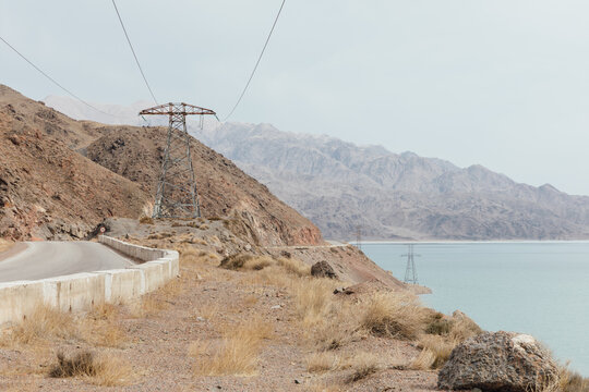 Scenic view of a winding mountain road with electricity pylons along the shore of Orto-Tokoy Reservoir, Issyk-Kul Region, Kyrgyzstan, on a hazy day