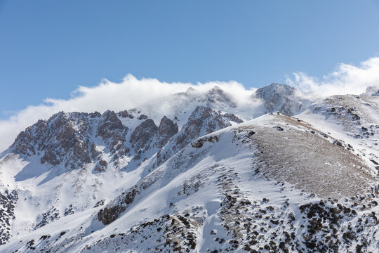 Snow-covered peaks and rocky ridgelines in the Alay Valley, Osh Region, Kyrgyzstan, with low clouds and clear blue sky, showcasing remote high-altitude nature