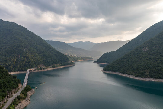 Wide landscape view of Piva Lake reservoir winding through a forested mountain canyon near Pluzine, Montenegro, with calm water, road bridge and dramatic clouds
