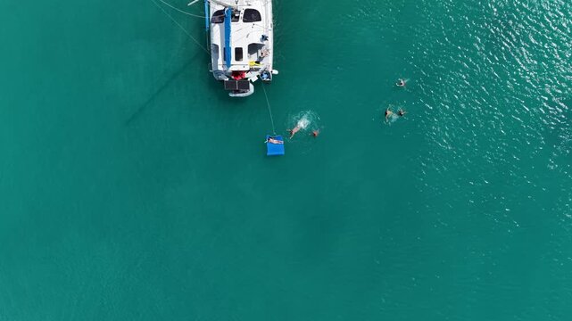 Aerial view of a luxury white catamaran anchored in the turquoise sea water of Saint Martin island in the Caribbean. People are swimming and relaxing on a blue floating mat next to the boat.
