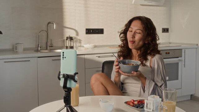 Asian young adult woman eating breakfast while vlogging with smartphone on tripod in kitchen. Clip is suitable for social media marketing, influencer lifestyle, and mobile technology ads