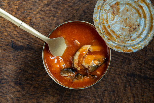 Top view of opened canned fish sprats in red tomato sauce with a golden fork on a dark wooden table, closeup