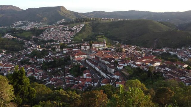 Aerial drone footage beginning with a couple overlooking Ouro Preto at sunset, then revealing the historic colonial city, iconic churches, traditional architecture, and surrounding mountains in golden