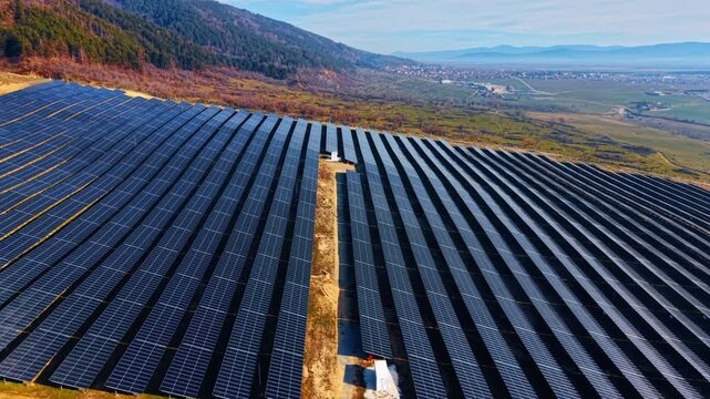 Parallel solar panel rows with central maintenance path. Drone view of dark modules, substation buildings and mountain foothills with autumn foliage.