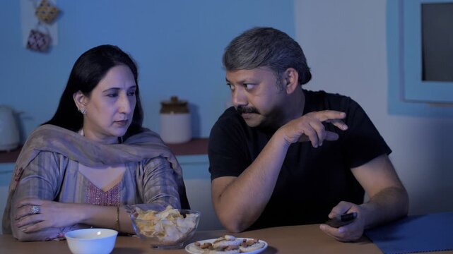 Mature couple enjoying their favorite TV program at home - leisure time. Medium shot of urban Indian husband-wife watching web-series and enjoying snacks late-night - happy marital life
