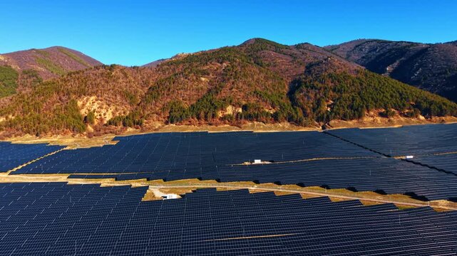 Sunlit solar farm covering hillside beneath forested peaks. Low angle aerial with dense panel coverage, clear blue sky and evergreen ridgeline.