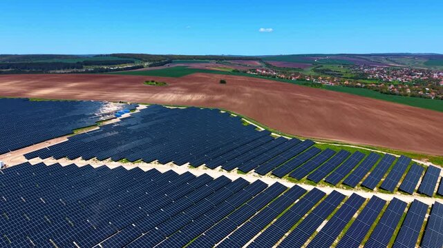 Solar power plant adjacent to farmland and rural village. Aerial view with dark panels, brown plowed fields and red rooftops under clear sky.