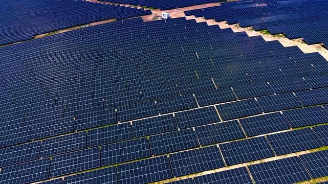 Aerial view of large solar panel array at industrial power plant. Overhead drone perspective of dark blue photovoltaic rows on sandy ground under hazy sky.