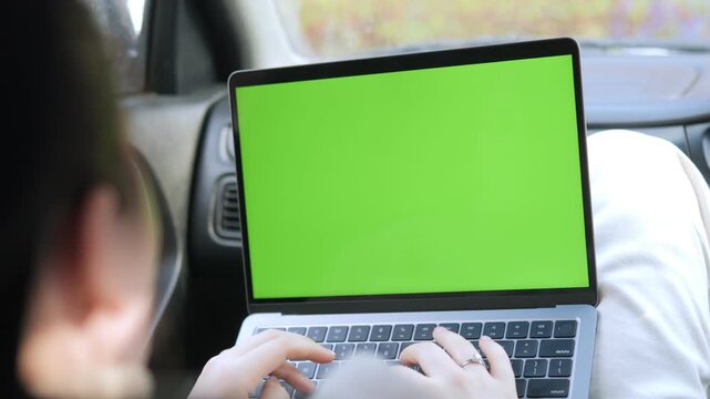 Over shoulder view of person typing on laptop with green screen inside car. Hands move quickly over keyboard. Focus on laptop screen and fingers. Person seems concentrated and focused
