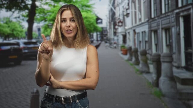 Woman with finger to lips for silence on a cobblestone street, arms crossed wearing white top and jeans, signaling shush to passerby; secretive hush.