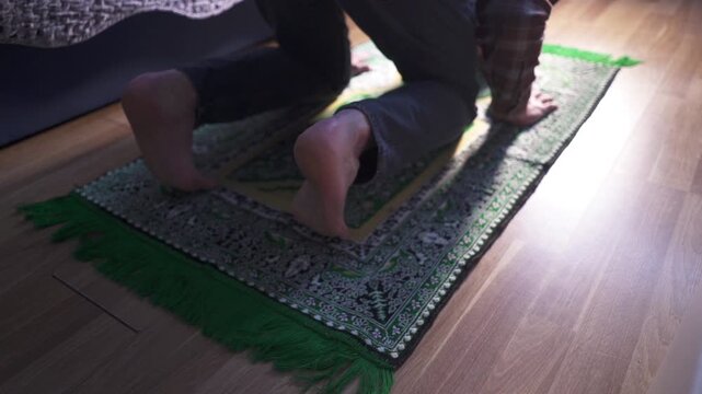 Muslim man performing prostration Sujood on a green prayer rug at home, bowing down in prayer.