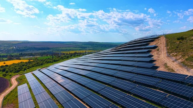 Large photovoltaic array on a hillside surrounded by farmland. Diagonal rows of dark panels climbing a green slope under a partly cloudy sky.