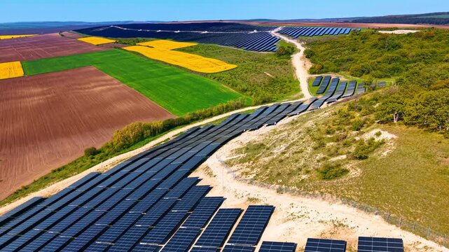 Elevated view of solar farm and colorful agricultural patchwork fields. Dark panel rows wind through sandy terrain beside green, yellow and brown crop parcels.