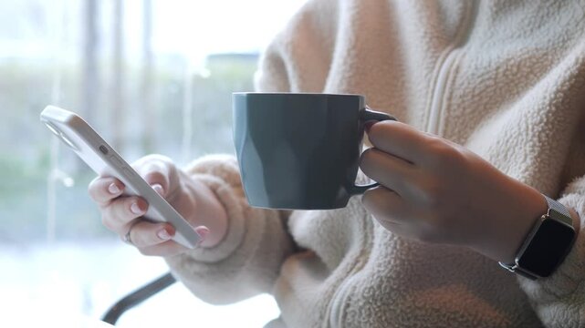Person holds phone and dark mug near window. Soft light illuminates cozy scene. Hands steady, focused on device screen. Comfortable indoor moment captured. Wristwatch visible on left arm