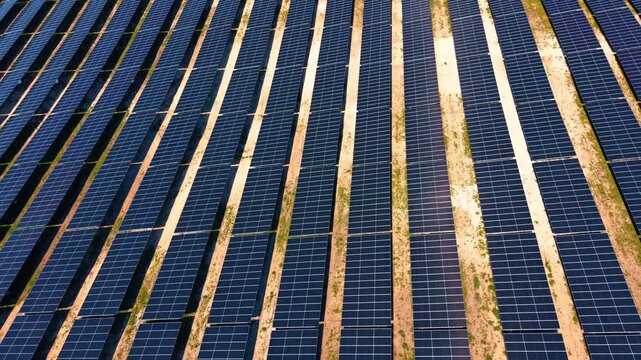 Oblique drone perspective of parallel solar panel rows at golden hour. Warm light casts long shadows across dark blue modules and sandy soil between rows.