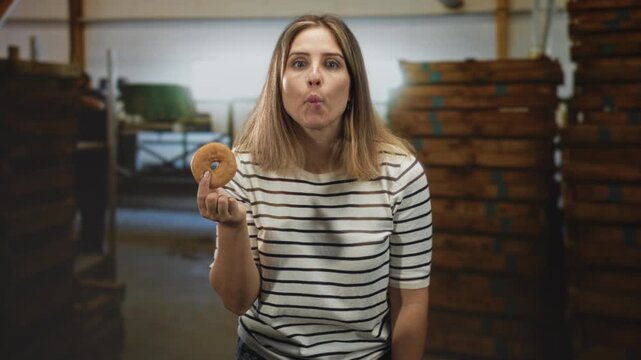 Woman holding a glazed donut in her hand, puckering lips and tilting head beside stacked wooden crates inside a building; playful curiosity.