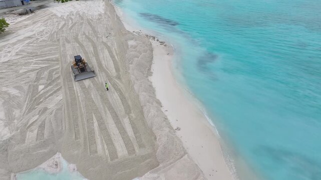 Aerial view of a bulldozer moving sand on a tropical beach with turquoise water and a worker standing nearby in Gulhi, Kaafu Atoll, Maldives.
