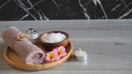 Luxury spa still life with white bath salt in a wooden bowl, a soft pink mauve towel tied with jute rope, a metallic cosmetic jar, pink frangipani flowers, and a candle on a light gray wooden table. P © kanjana