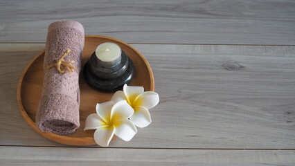 Minimalist zen spa still life with a soft mauve pink towel tied with rope, a scented candle on stacked black massage stones, and white frangipani flowers in a circular wooden tray. Set on a light gray © kanjana