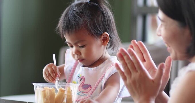 Child feeding snack independently, A little child feeds itself, while mother claps her hands, expressing family love, learning, care, and warm bonding moments, Thai Asian